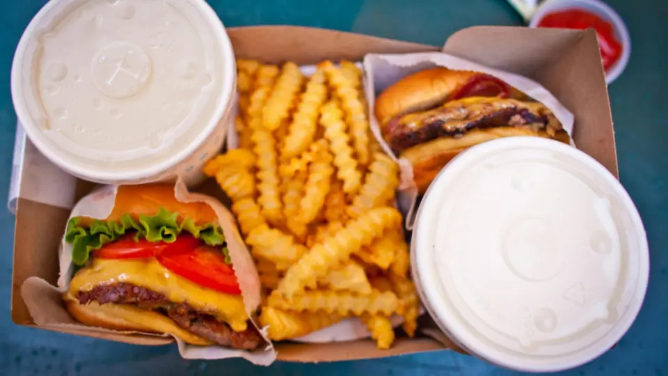 A tray of fast food is displayed from above. It includes two cheeseburgers with lettuce, tomato, and melted cheese, a large portion of crinkle-cut fries in the center, and two takeaway cups with lids.