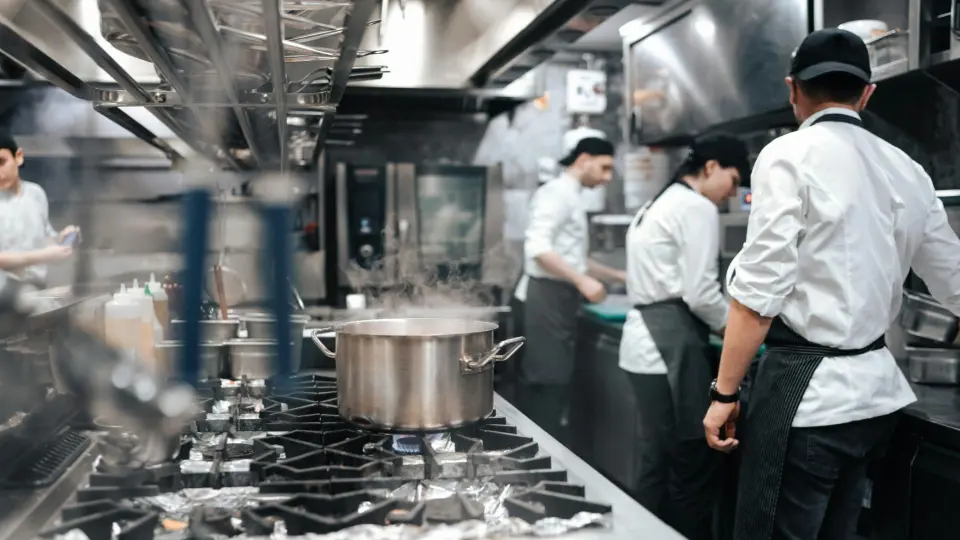 A group of chefs in professional white uniforms working in a high-speed commercial kitchen, focused on food preparation and plating at a stainless steel station.