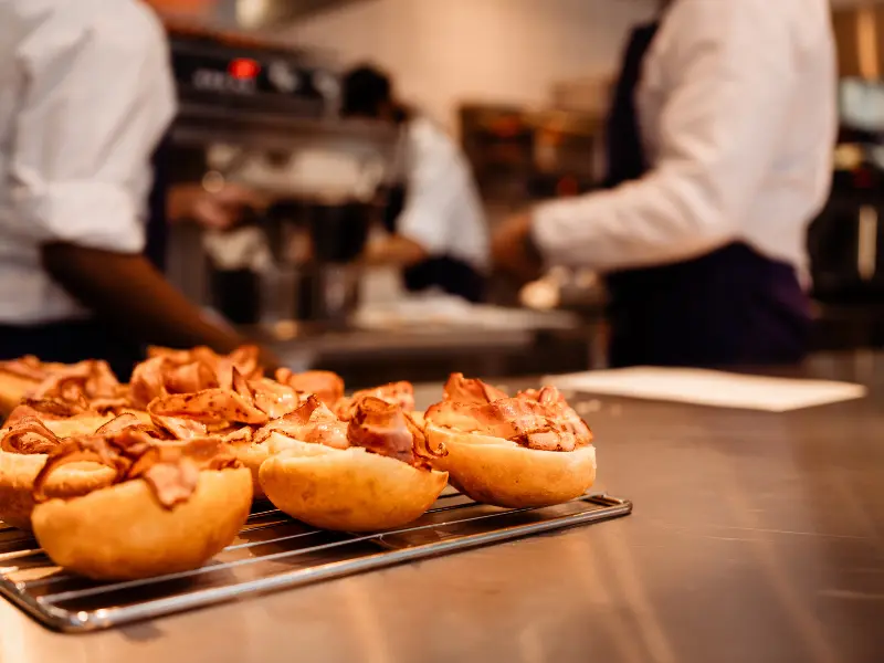 The image shows freshly prepared sandwiches with slices of cooked meat placed on a cooling rack in a professional kitchen.