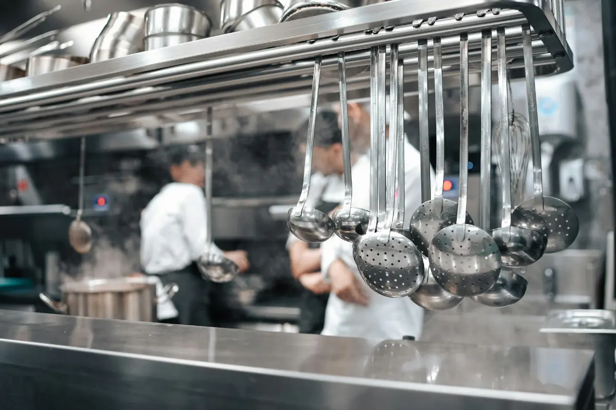The image shows a professional industrial kitchen. In the foreground, there is a row of stainless steel ladles and cooking utensils hanging from a metal rack above a counter.