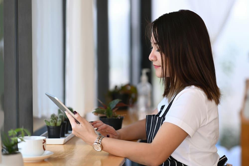 A person wearing a white shirt and a black and white striped apron sits at a wooden counter by a window, using a tablet.