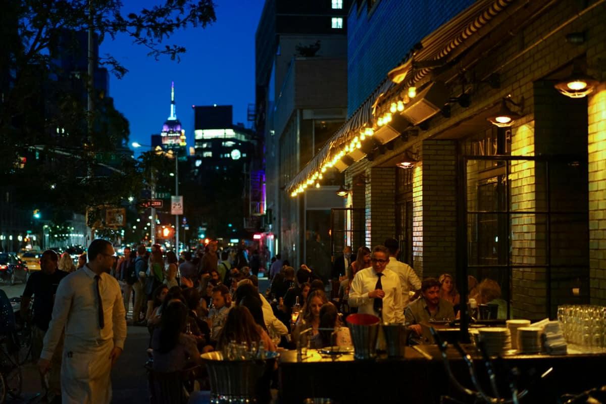 Outdoor dining scene at night in one of the best neighborhoods in New York for food delivery, full of people and city lights.