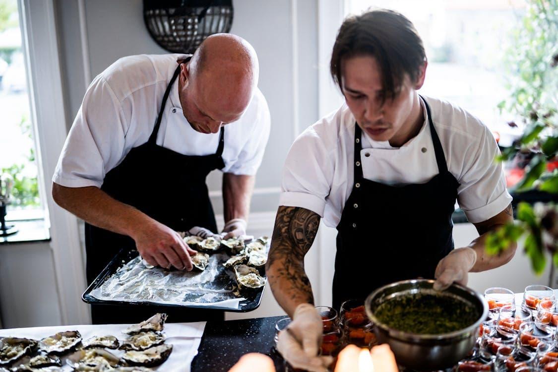 The image shows two chefs working in a kitchen, carefully preparing food. One of the chefs, bald and focused, is arranging oysters on a tray, while the other chef, with a tattoo on his arm, is holding a bowl and seemingly working with some green sauce or seasoning. Both are wearing white chef's uniforms with black aprons and are intensely focused on their tasks, suggesting a professional kitchen setting, possibly for catering or fine dining. The workspace is well-lit, and the chefs' actions appear coordinated.