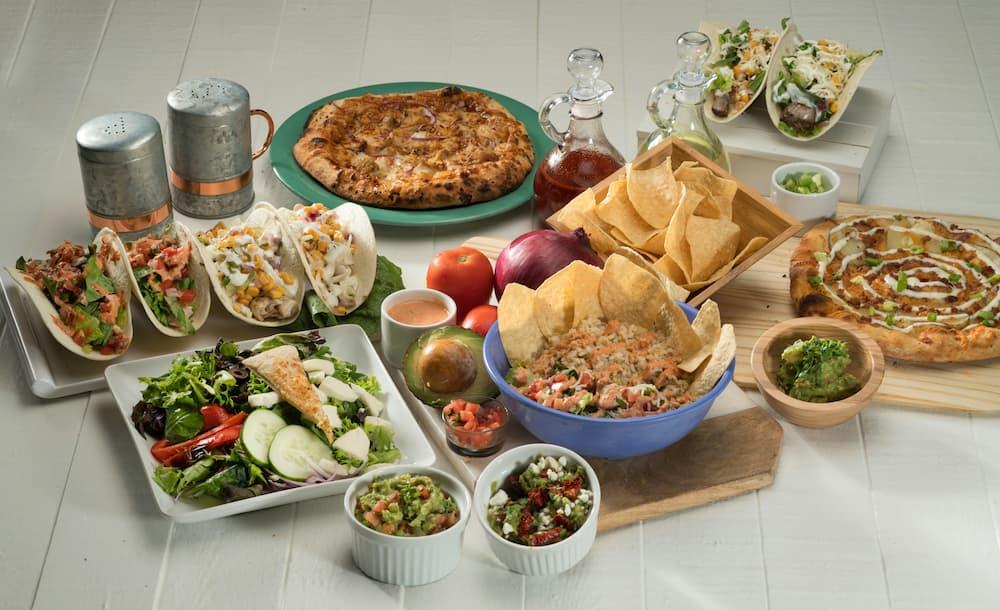 Cinco de Mayo party spread showcasing vibrant Mexican dishes: tacos stuffed with fresh vegetables, bowls of guacamole and salsa, rice garnished with tomatoes and herbs, crispy tortilla chips, and pizzas with creative toppings. The table features festive touches including metal shakers, sauce bottles, and colorful produce like tomatoes and avocados, all set on a clean white surface for a lively celebration.