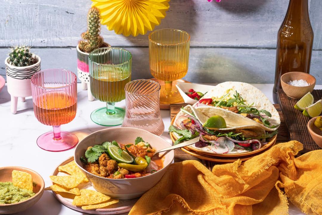 Cinco de Mayo celebration setup featuring a table adorned with festive elements: colorful drinks, a bowl of Mexican cuisine garnished with lime and cilantro, tortilla chips with guacamole, and a plate of tacos accompanied by fresh vegetables and lime.