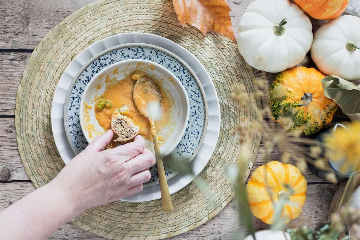 Close-up of a bowl of creamy pumpkin soup served with a piece of bread, surrounded by decorative pumpkins on a rustic table setting.