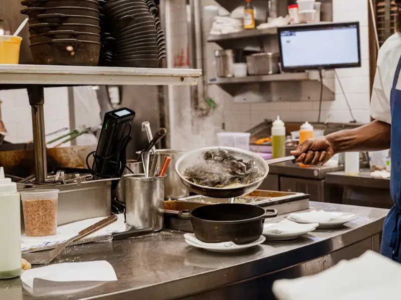 Chef cooking fish in a busy commercial space with a kitchen, surrounded by utensils, plates, and cooking equipment.