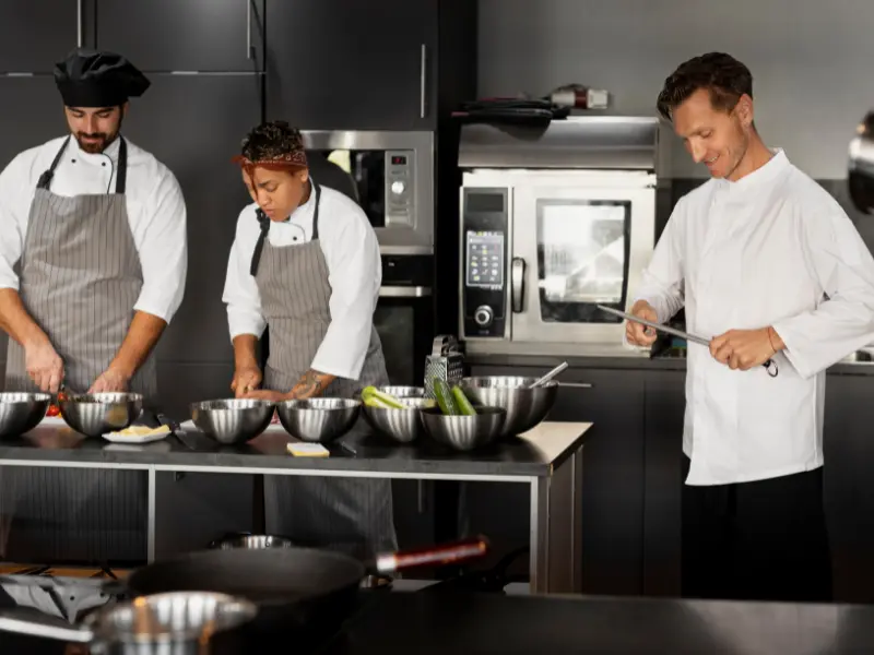 The image depicts a scene of teamwork and preparation inside a modern, professional kitchen.