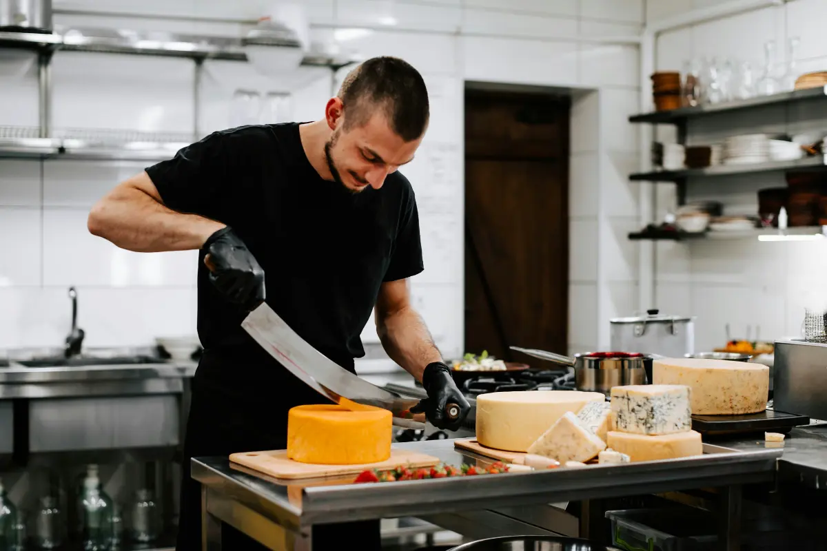 Man slicing cheese in a clean commercial space with kitchen, featuring stainless steel surfaces and organized shelves.