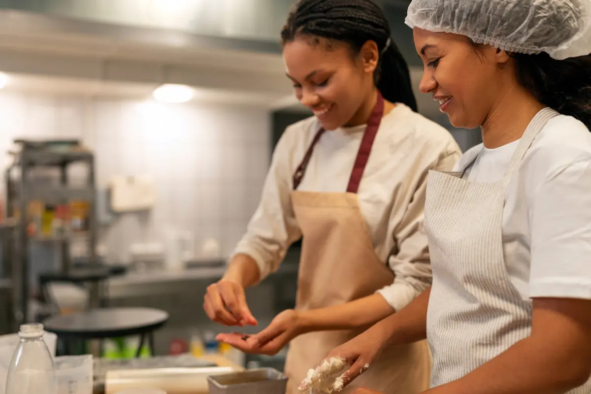 The image depicts two women working together in what appears to be a commercial or professional kitchen setting, possibly within a culinary incubator.