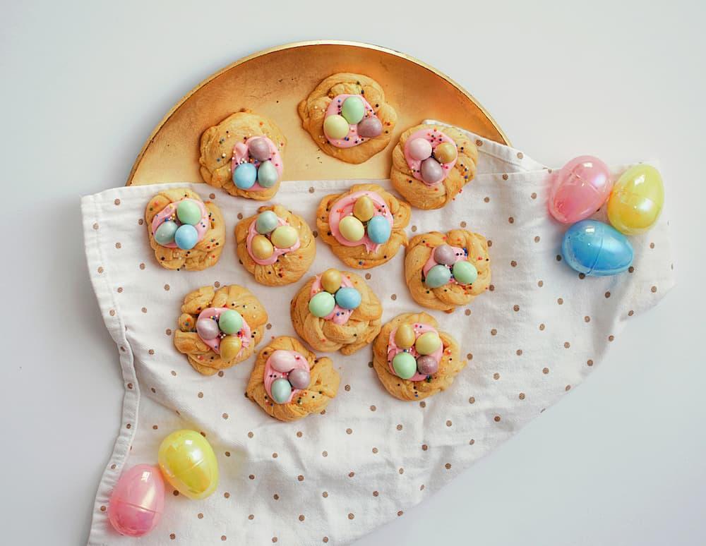 Easter desserts on display, featuring cookies adorned with colorful candy eggs. The cookies are arranged beautifully on a golden tray atop a white cloth decorated with gold polka dots. Surrounding the tray are vibrant plastic Easter eggs in pink, yellow, and blue hues, adding a festive touch.