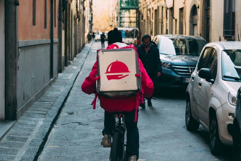 App delivery person on a bike making a delivery on a busy street. They are wearing a red thermal backpack with a food symbol.