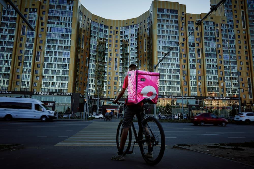 Food delivery person with a bright pink backpack is standing next to a bicycle in front of a modern yellow and gray apartment building.