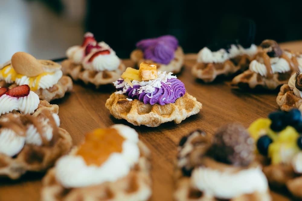 A beautifully presented assortment of waffles laid out on a wooden surface, showcasing creative food plating and presentation. Each waffle features a unique combination of toppings, including whipped cream, vibrant fruits, colorful creams, and garnishes, highlighting culinary artistry and attention to detail.