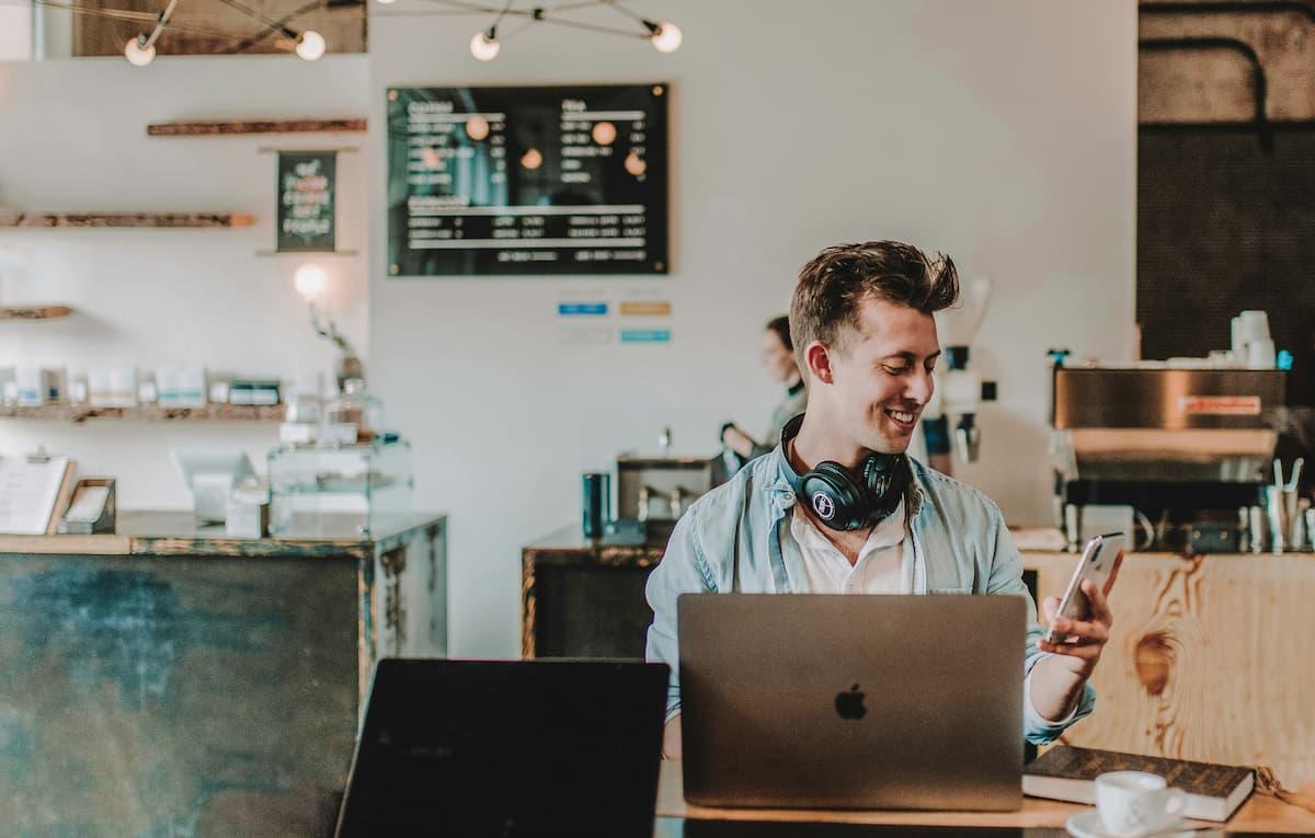 Smiling man wearing headphones, working on a laptop and checking his phone in a modern coffee shop with a menu and machines in the background.