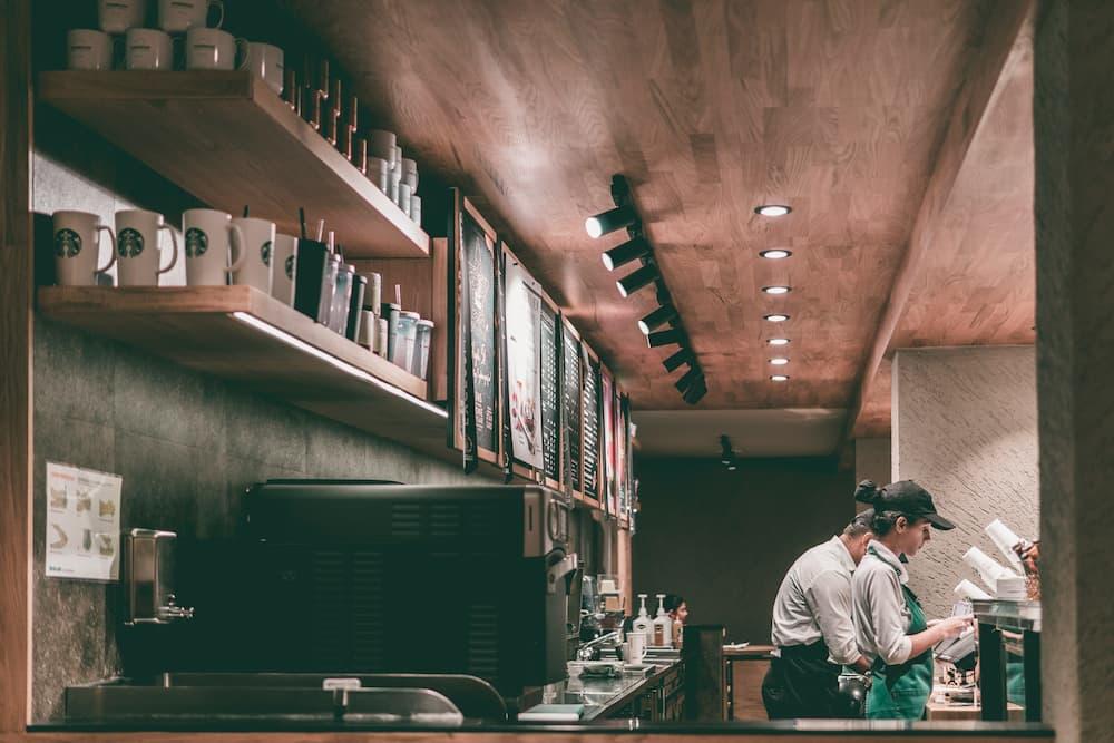 A modern coffee shop environment, with baristas working at a counter, shelves with cups, and a menu visible on the wall