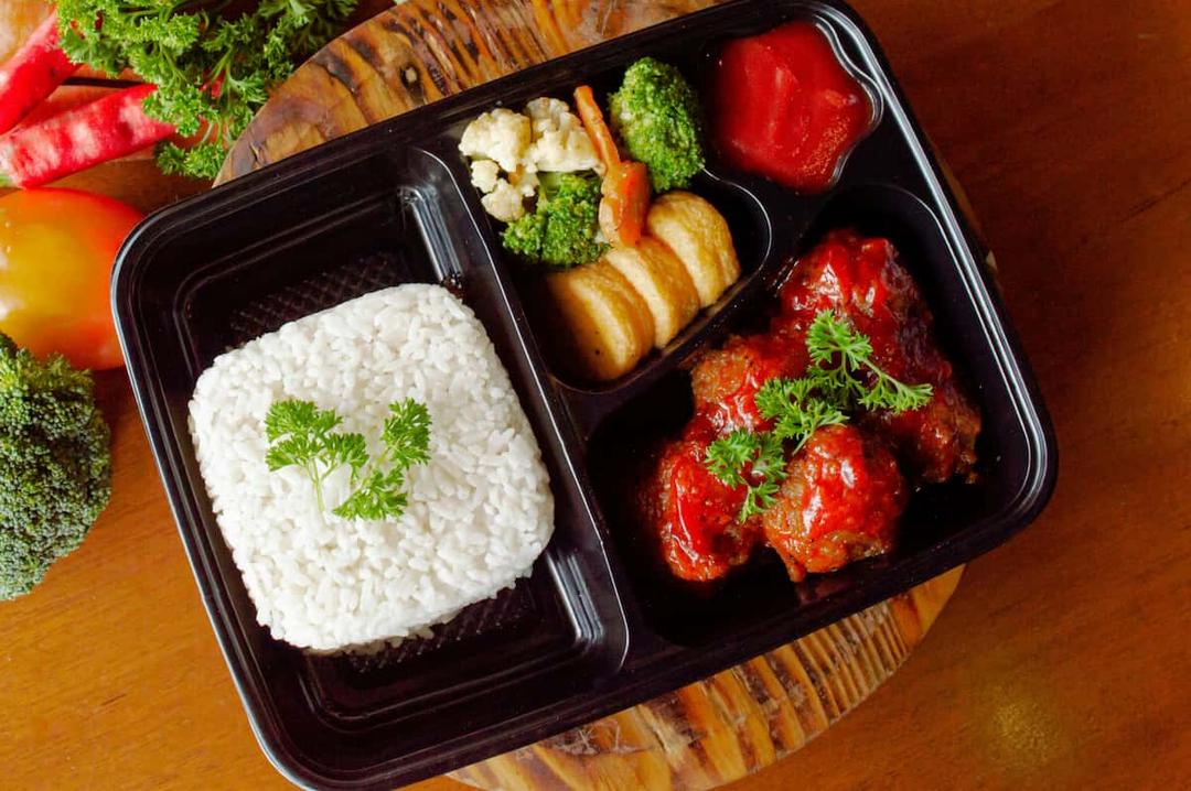 A neatly arranged frozen meal in a black tray, featuring white rice garnished with parsley, steamed vegetables, and saucy meatballs, placed on a wooden table with fresh vegetables in the background.