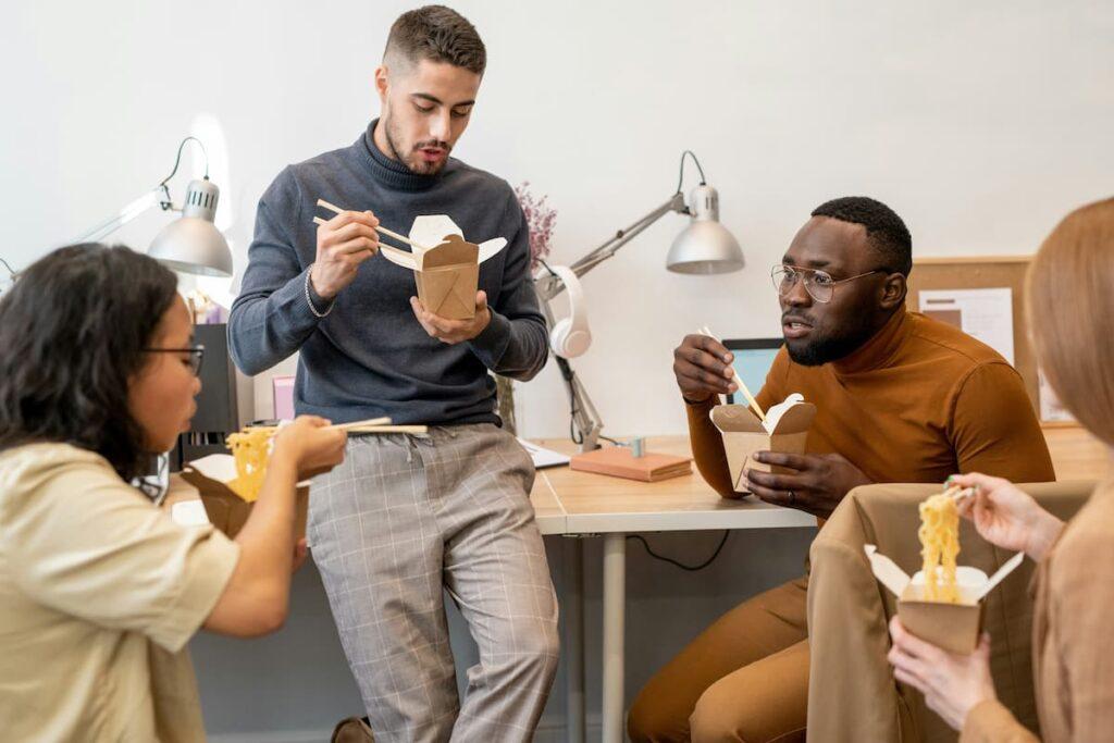 A group of young people sharing a meal with Asian food in typical packaging, in a modern office setting.