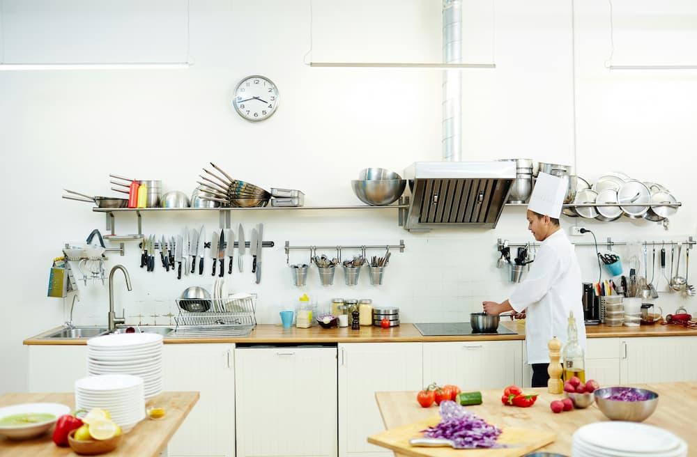 A professional kitchen with a chef cooking at a stovetop. The kitchen is well-organized with various utensils, knives, and pots hanging on the wall.