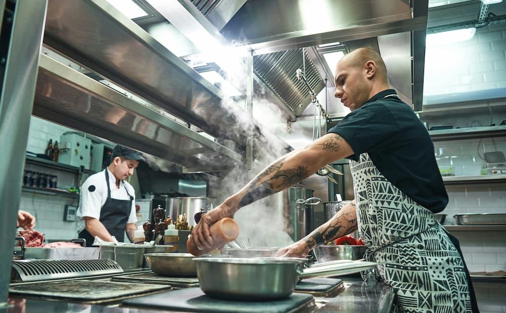 A bustling commercial kitchen where two chefs are at work. In the foreground, a chef wearing a patterned apron and black shirt is seasoning food in a steaming pan on the stove, showcasing their tattooed arms.