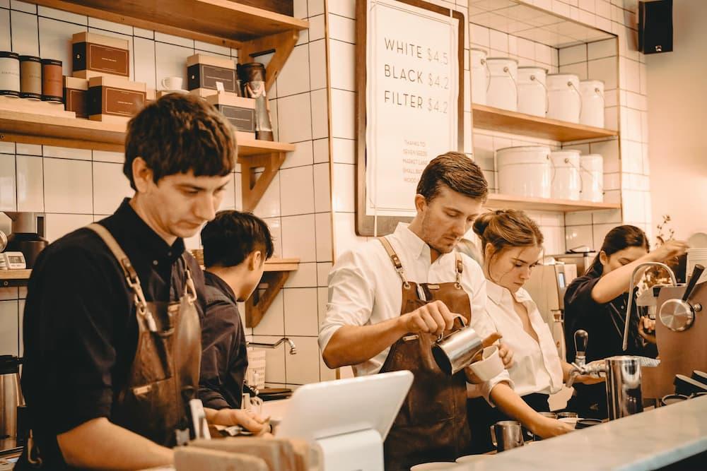 A group of baristas working behind the counter in a coffee shop. They are wearing aprons and are engaged in various tasks such as making coffee and handling equipment.