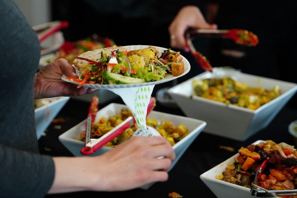 person holding a paper plate filled with a variety of colorful food items, including salad, vegetables, and protein, while standing in front of a catering table with several white square bowls containing different types of food.