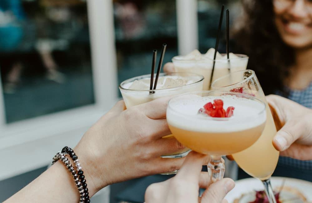 A group of people raising their cocktail glasses in a celebratory toast. The drinks are varied, with some garnished with fruit and others with straws, capturing the festive atmosphere of a cocktail hour.