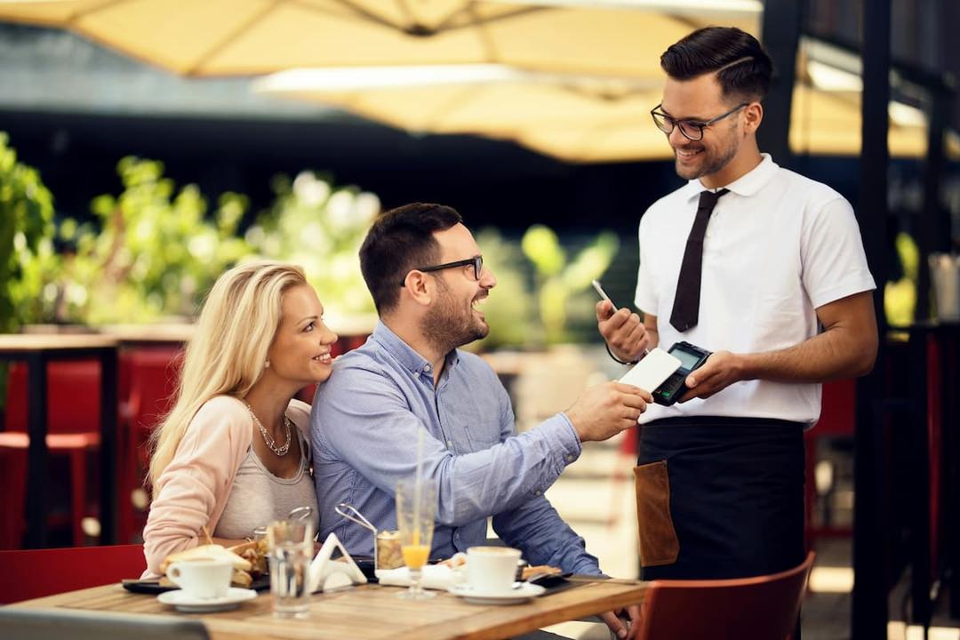 Smiling couple at an outdoor restaurant paying with a contactless card while a waiter holds the card reader.