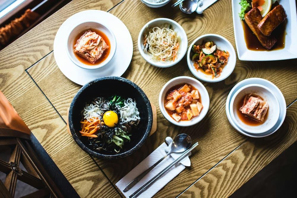 A top-down view of a table featuring a traditional Korean meal. The centerpiece is a bowl of bibimbap with a raw egg yolk on top, surrounded by colorful vegetables, rice, and seaweed