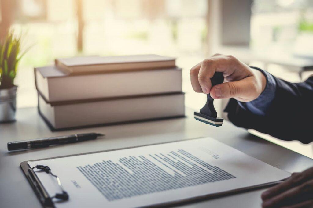 Person stamping a document on a desk, with books and a pen in the background, symbolizing approval or certification in a professional setting.