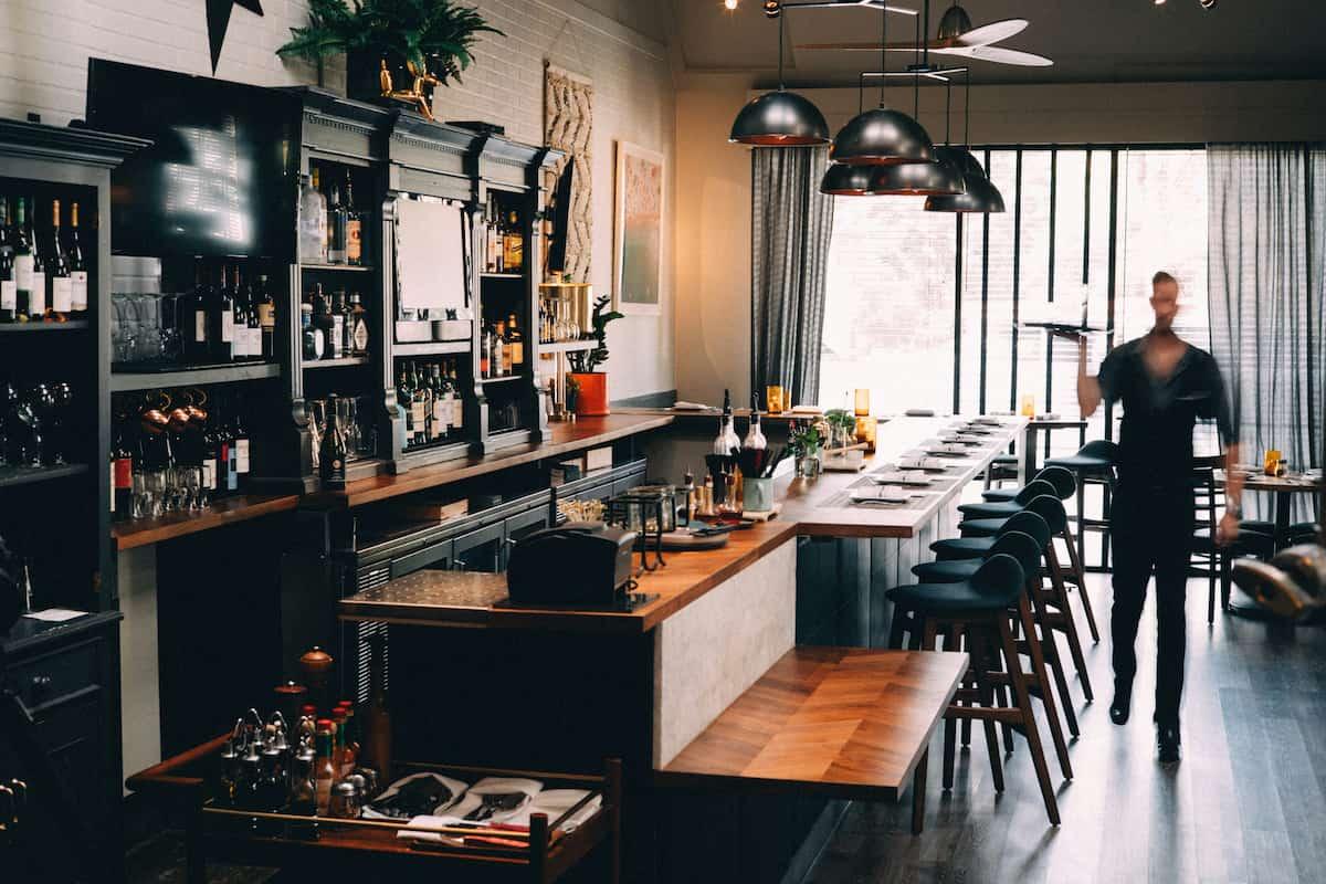 A stylish bar and restaurant interior featuring a wooden countertop, shelves stocked with bottles, and modern pendant lighting. A server is seen walking in the background, adding a lively touch to the elegant and cozy ambiance.