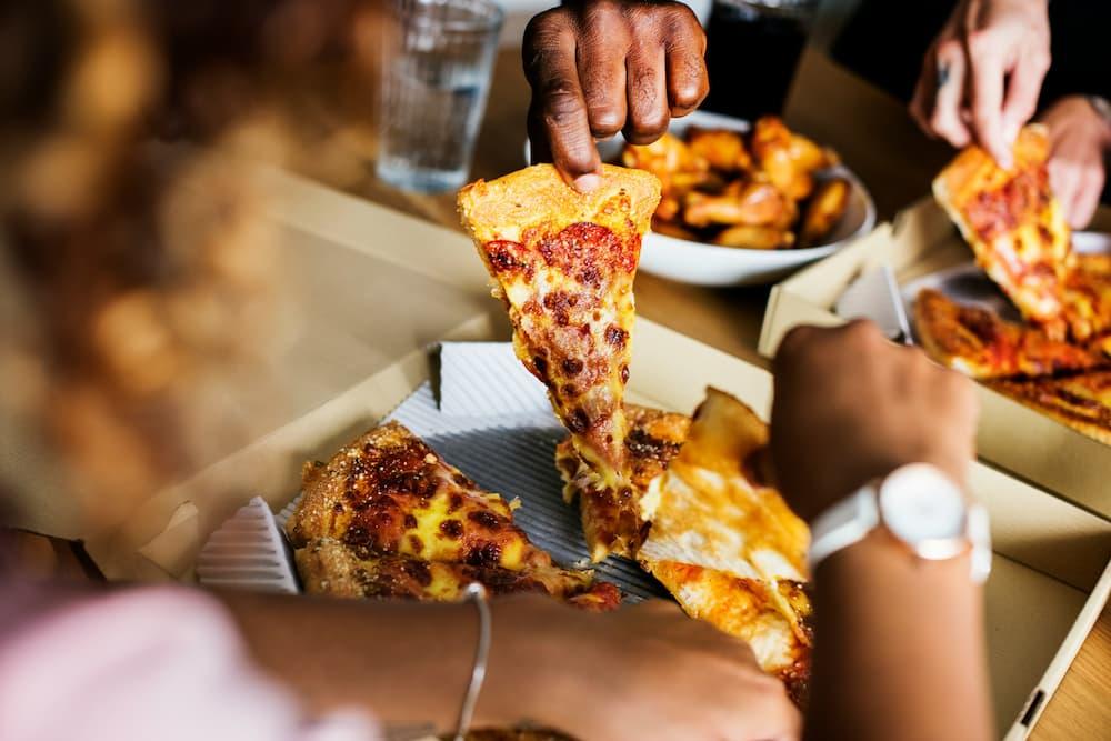 A close-up view of a group of people sharing a pizza. One person lifts a slice of pepperoni pizza topped with melted cheese from the box, while others reach for slices.