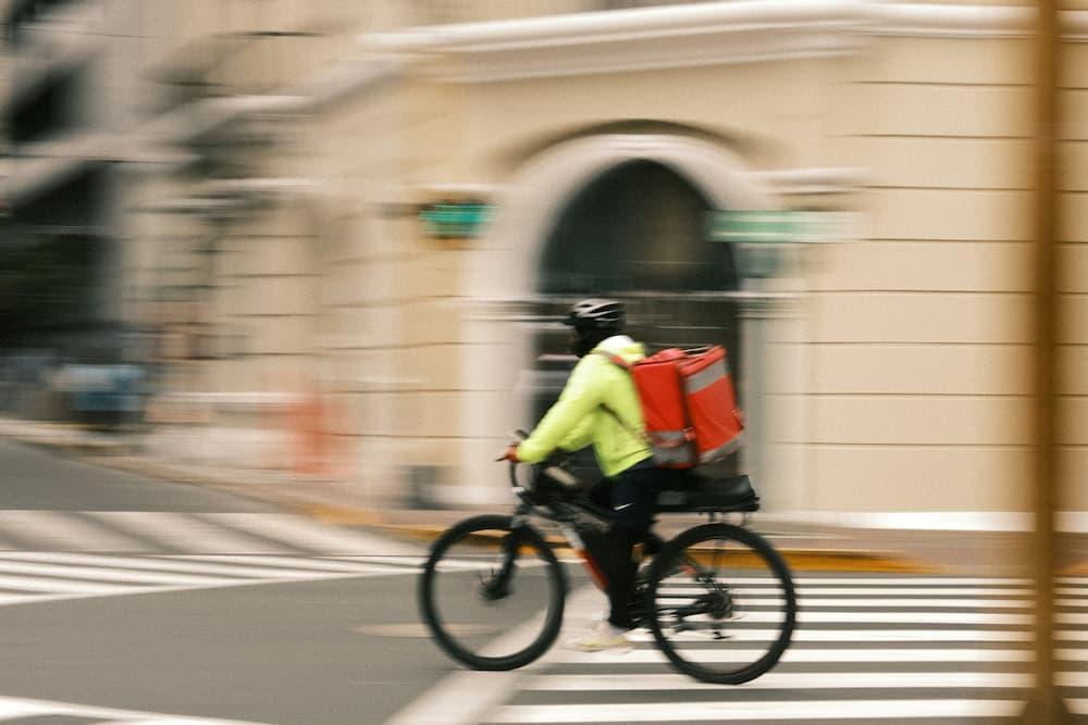 "A person wearing a bright yellow-green jacket and a black helmet is riding a bicycle across a crosswalk in an urban area. The cyclist has a large red insulated delivery bag on the back, indicating that the person is likely a food delivery worker