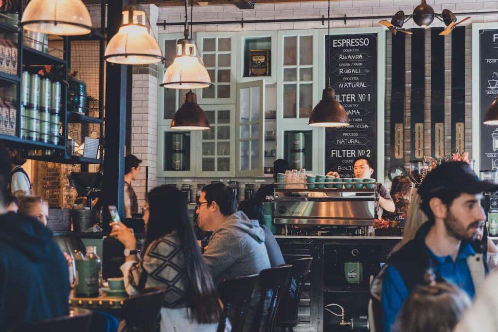 Customers enjoying coffee and meals in a cozy, well-lit cafe with a barista preparing drinks in the background.