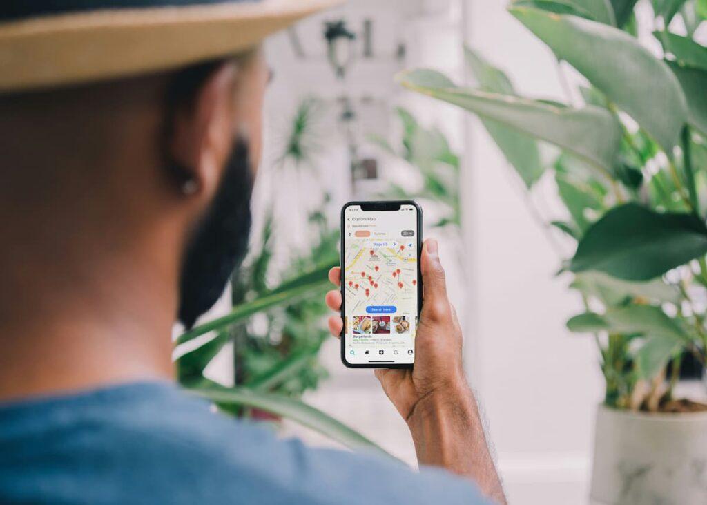 A man holding a smartphone with a restaurant map and location markers on the screen, possibly searching for nearby dining options using a food delivery or restaurant discovery app. Surroundings include plants and a casual setting.