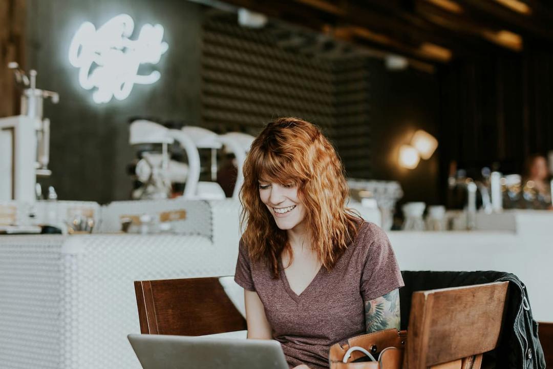A smiling woman with red hair using a laptop in a modern coffee shop, with blurred barista equipment in the background.