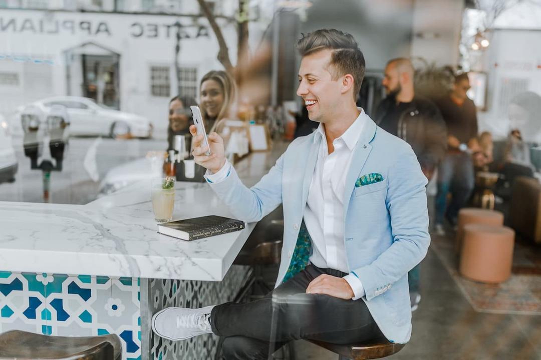 Smiling man in a light blue blazer sitting at a modern café, holding a smartphone and interacting with an app, representing customer engagement and loyalty in a restaurant setting.