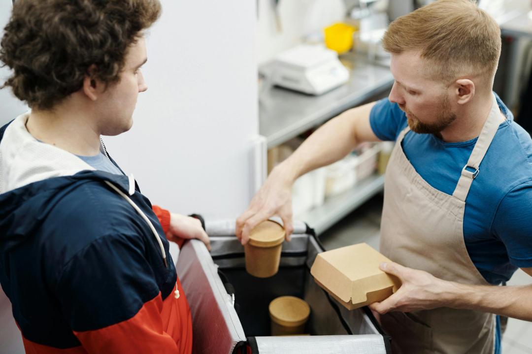 The image shows a restaurant employee or chef wearing a beige apron and a blue t-shirt. He is carefully placing eco-friendly cardboard containers into a large, insulated delivery thermal bag.