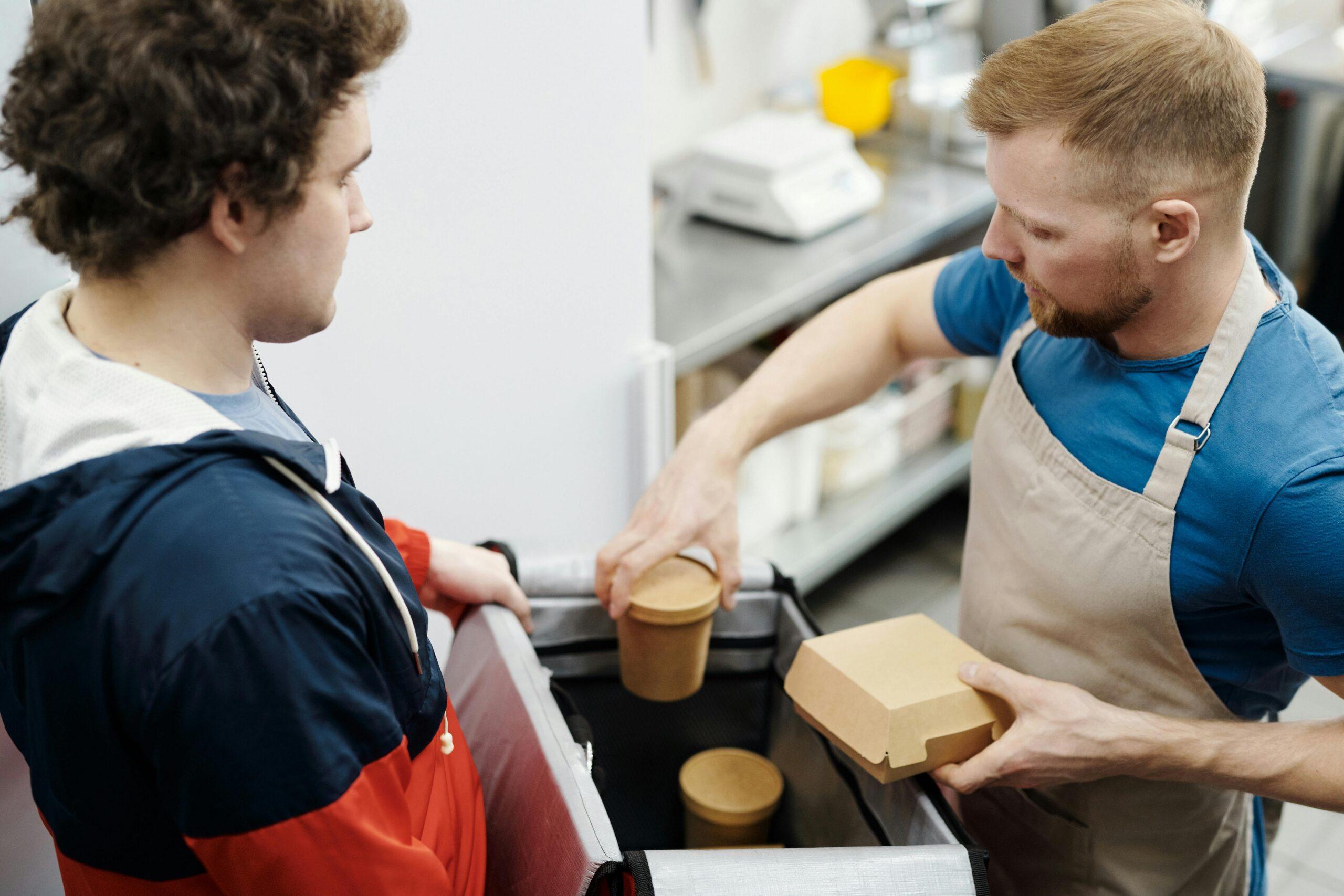 The image shows a restaurant employee or chef wearing a beige apron and a blue t-shirt. He is carefully placing eco-friendly cardboard containers into a large, insulated delivery thermal bag.