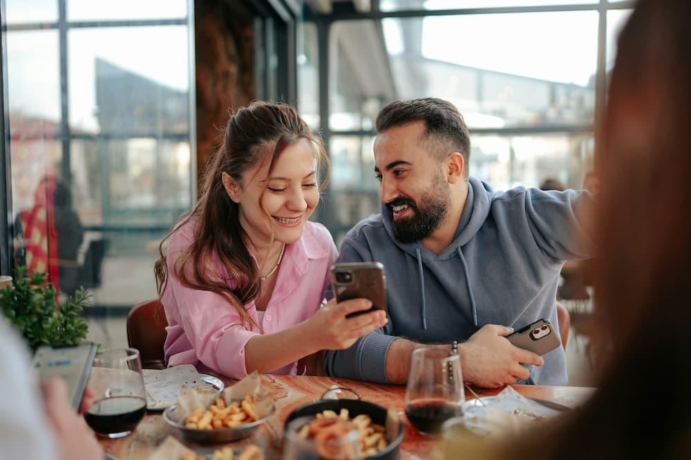 A couple having fun while looking at something on their phones in a modern café, with dishes and drinks on the table
