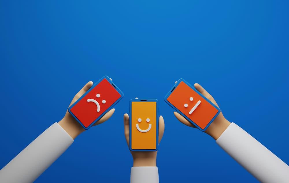 Three hands holding smartphones with different emojis: one sad, one happy, and one neutral, on a blue background