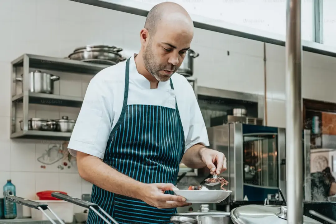 A chef is working in a commercial kitchen. He is wearing a white chef's coat and a dark apron with blue stripes. He appears focused as he carefully plates food using a spoon. The kitchen around him is clean and organized, with metal shelves holding various pots and pans, and a stove or counter with cooking equipment in the foreground. Stainless steel surfaces and utensils are visible, indicating a professional cooking environment.