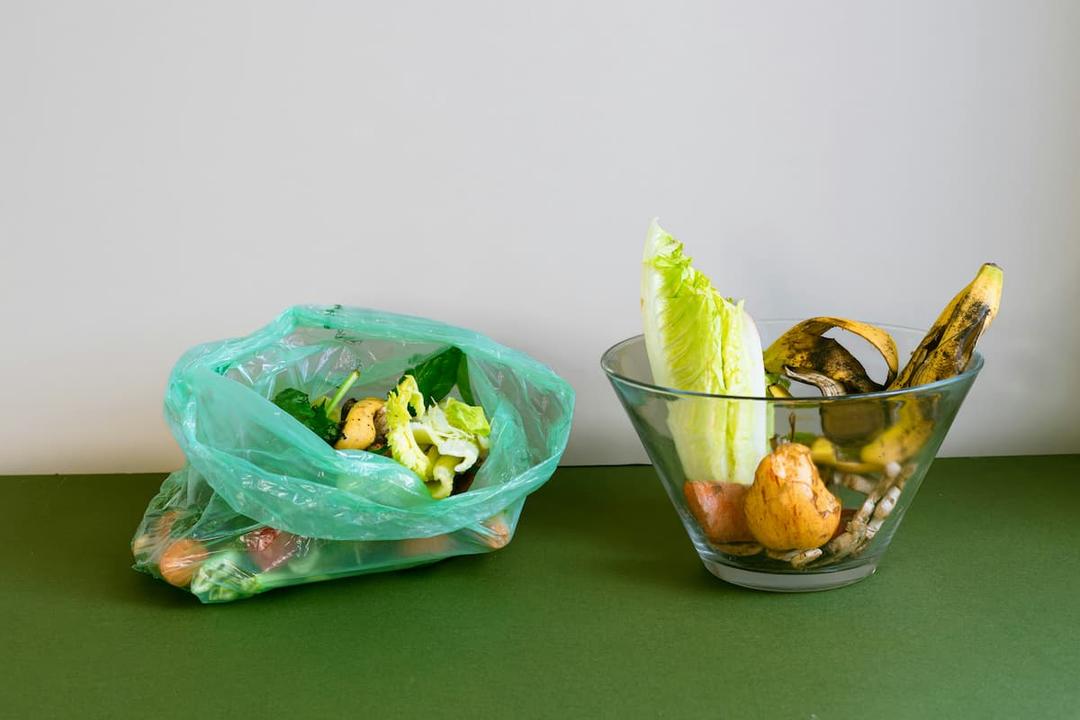 Organic waste separation: a green plastic bag filled with vegetable scraps on the left and a glass bowl with food waste, including lettuce, bananas, and fruits, on the right.